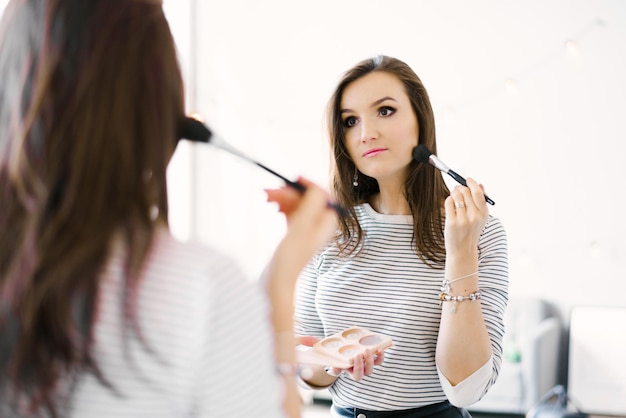 Schönes Mädchen trägt mit einem Make-up-Pinsel Rouge auf ihr Gesicht auf, während sie zu Hause in einem Zimmer steht und in den Spiegel schaut. Ein kaukasisches Mädchen in einem gestreiften Hemd macht ihr eigenes Make-up