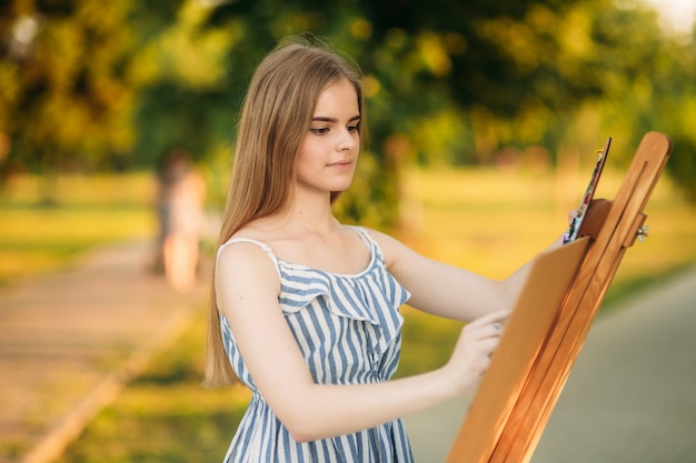 Schönes Mädchen zeichnet ein Bild im Park unter Verwendung einer Palette mit Farben und einem Spatel. Staffelei und Leinwand mit einem Bild.