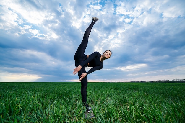 Schönes Mädchenmodell auf grünem Gras tun Yoga. Eine schöne Brünette auf einem grünen Rasen führt akrobatische Elemente durch. flexible Turnerin in schwarz macht Übungen