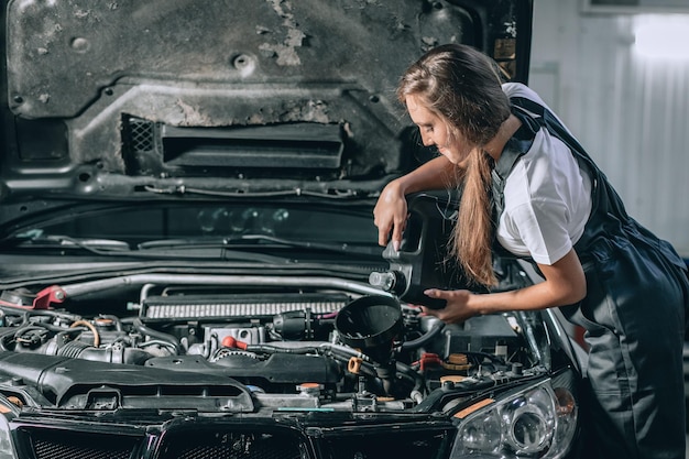 Schönes Mechanikermädchen in einem schwarzen Overall und einem weißen T-Shirt wechselt das Öl in einem schwarzen Auto. Autoreparaturkonzept