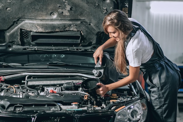 Schönes Mechanikermädchen in einem schwarzen Overall und einem weißen T-Shirt wechselt das Öl in einem schwarzen Auto. Autoreparaturkonzept