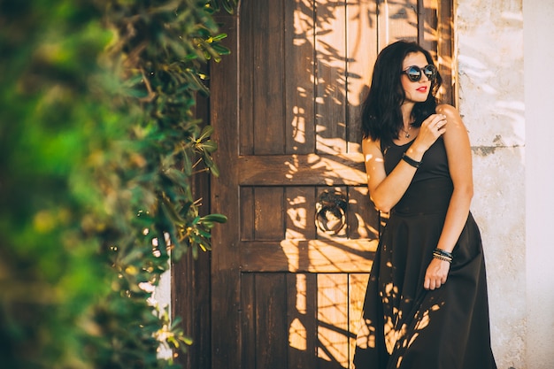 schönes modernes junges Brunettemädchen in der Sonnenbrille und in einem langen schwarzen Kleid, das an einer Holztür steht