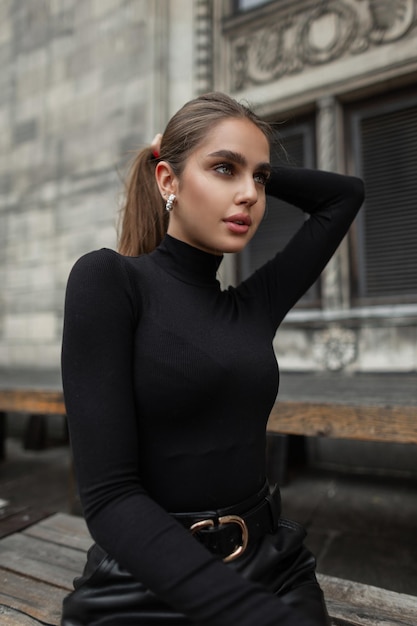 Schönes, modisches, elegantes, frisches Mädchenmodell mit Ohrringen in trendigen schwarzen Kleidern sitzt in der Stadt Urbanes weibliches Porträt einer hübschen Frau