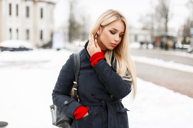 Schönes modisches junges Mädchen in einem stilvollen Modemantel mit einer Tasche, die in der Stadt an einem Wintertag geht