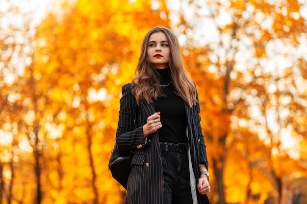 Schönes modisches Mädchen mit roten Lippen in modischer Kleidung mit Blazer und Pullover mit Lederrucksack geht bei Sonnenuntergang im Park mit orangefarbenen Blättern