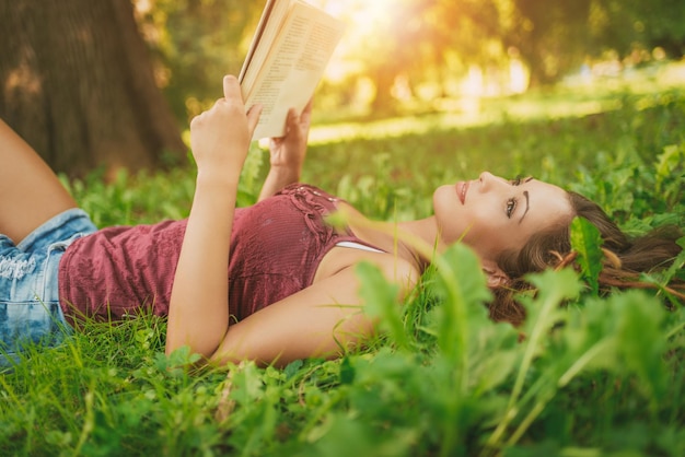 Schönes nachdenkliches Mädchen, das im Gras liegt und Buch liest.