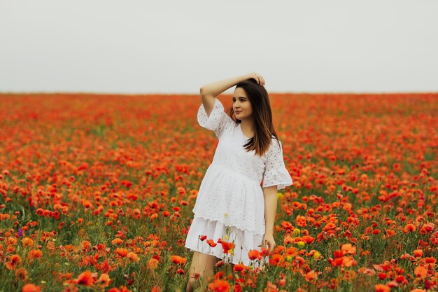 Schönes niedliches brünettes Mädchen im Mohnfeld am Sommertag in einem weißen Kleid