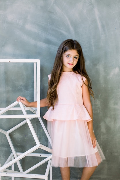 Schönes niedliches kleines brünettes Mädchen in einem hellen Kleid, das auf einer grauen Wand steht