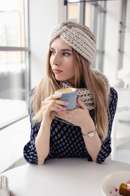 Schönes niedliches Wintermädchen, das Strickmütze und Handschuhe mit Kaffeetasse trägt. Attraktives Mädchen mit heißem Getränk auf Caféhintergrund. Kaffee trinken und das Leben genießen.