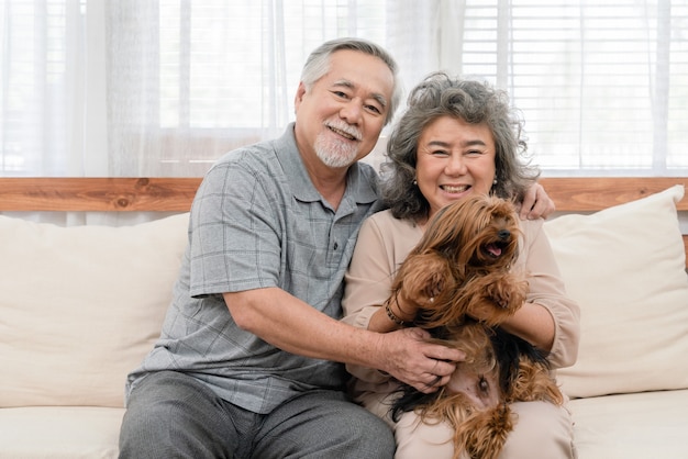 Schönes Paar asiatischer älterer Menschen mit ihrem Hund, der zu Hause auf dem Sofa sitzt