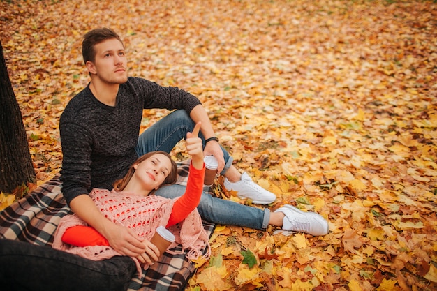 Schönes Paar, das allein im Herbstpark sitzt und liegt. Sie schauen in die gleiche Richtung. Frau, die auf den Beinen des Kerls liegt. Guy sitzt auf einer Decke. Sie halten eine Tasse Kaffee.