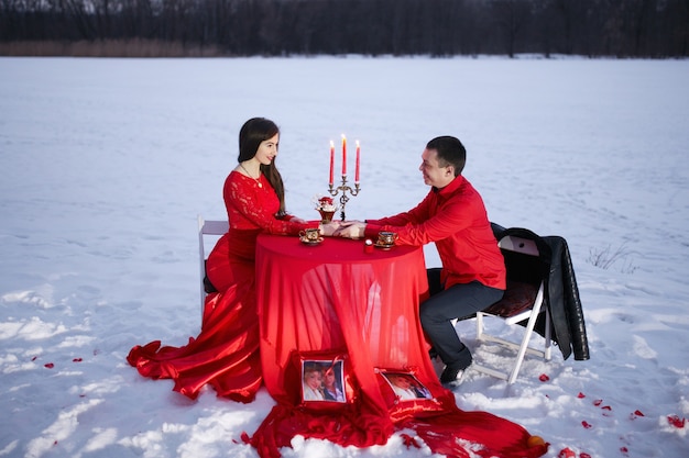 Schönes Paar, das an einem Tisch mit einem Kerzenhalter auf einem Hintergrund des Schnees in den roten Kleidern sitzt