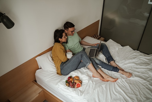Schönes Paar, das einen Film mit romantischen Gefühlen sieht, auf dem Bett mit Kaffee und einem Teller mit Croissants und frischen Früchten sitzt. Glückliche Familie, die in eine neue Wohnung zieht.
