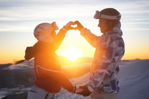 Schönes Paar Händchen haltend in Herzform auf schneebedecktem Gipfel bei Sonnenuntergang. Winterurlaub