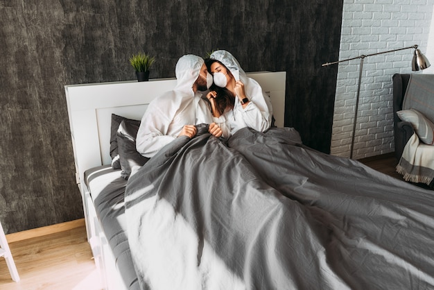 Schönes Paar in weißen Schutzkostümen mit Kapuzen und Gesichtsmasken zu Hause auf dem Bett