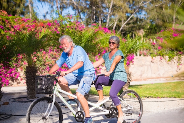 Schönes Paar von Senioren und Älteren verheiratet, die ein Doppelfahrrad fahren, Tandem, zusammen, um Spaß mit einem großartigen und sonnigen Tag zu haben - Frau und Mann