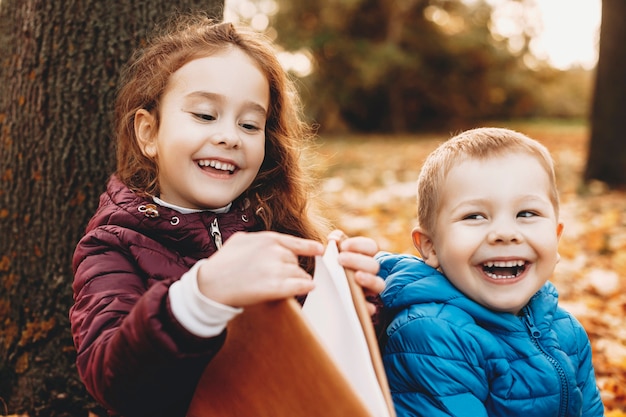 Schönes Porträt eines niedlichen kleinen Bruders und einer Schwester, die Spaß am Lachen haben, während Mädchen ein Buch öffnet, während Junge draußen im Park wegschaut.