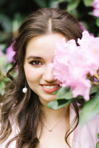 Schönes Porträt eines reizenden Mädchens, das durch blühenden Rhododendron der lila rosa Blumen im Frühjahr im Garten umgeben steht. . Professionelles Make-up. Eine Blume mit geschlossenen Augen, lächelt sie