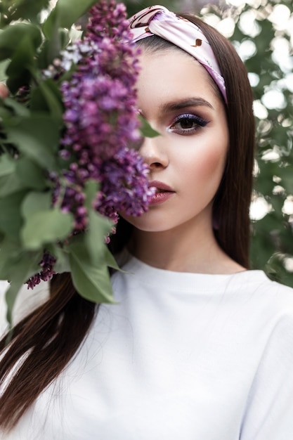 Schönes Porträt schöne Frau mit Farb-Make-up im Vintage-Bandana im weißen trendigen Hemd mit lila Blumen im Park. Glamouröses Mädchen hält luxuriöse Blumenstraußflieder in der Nähe des Gesichts auf Hintergrundblättern.