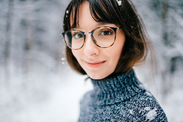 Schönes positives Brunettemädchen in den modischen Gläsern, die Kamera betrachten. mit schnee auf den haaren.