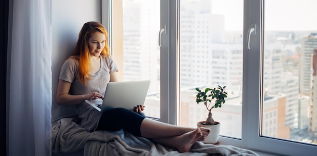 Schönes positives Mädchen mit Laptop, sitzend auf der Fensterbank in der Stadtwohnung. Junge rothaarige Frau, die zu Hause arbeitet. Freiberufliches Konzept.