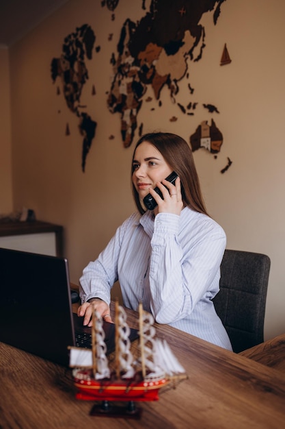 Schönes Reisebüro spricht am Telefon, schaut in die Kamera und lächelt, während sie am Tisch im Büro sitzt