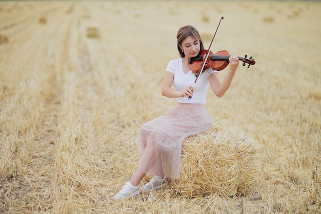 Schönes romantisches Mädchen mit losen Haaren, die Geige auf dem Feld nach der Ernte spielen