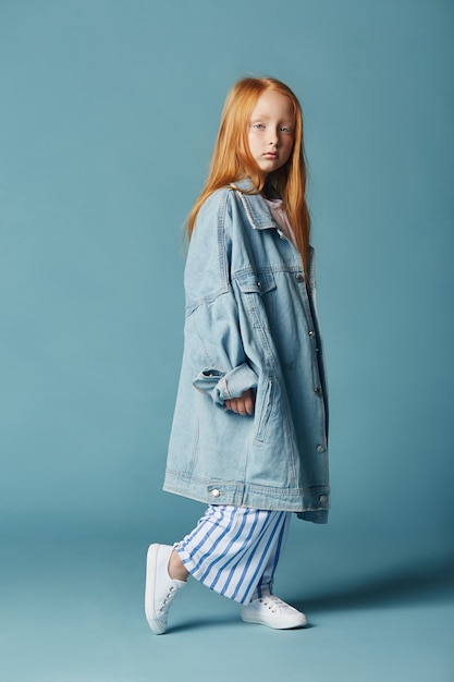 Schönes rothaariges Baby mit langen Haaren in einer großen blauen langen Jeansjacke.