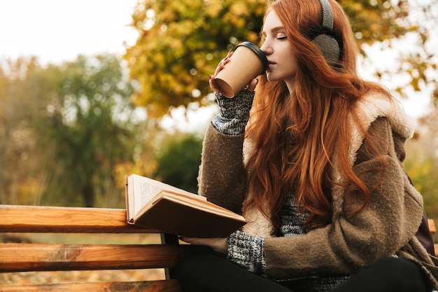 Schönes rothaariges junges mädchen, das musik mit kopfhörern hört, während auf einer bank sitzt, ein buch liest, kaffee trinkt