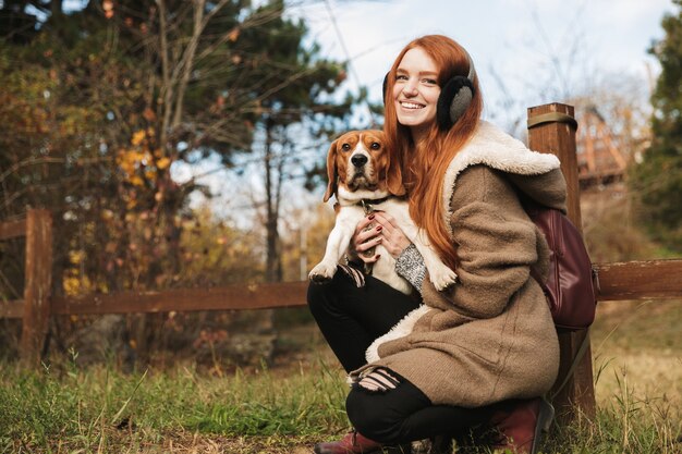 Schönes rothaariges junges Mädchen, das Musik mit Kopfhörern hört, während sie mit ihrem Hund im Park spielt