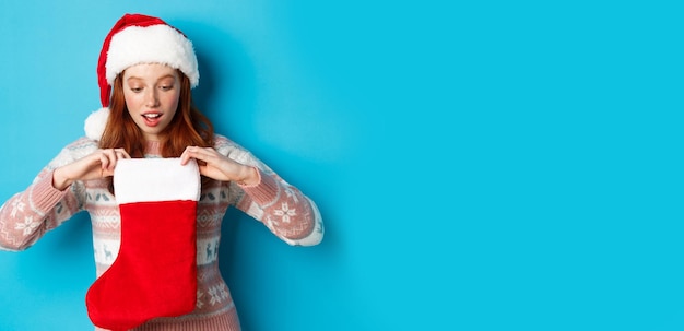 Schönes rothaariges Mädchen in Sankt-Hut öffnet Weihnachtsstrumpf und sieht überrascht aus, Weihnachts-GIF zu erhalten