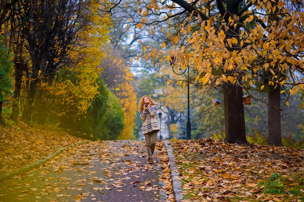 Schönes rothaariges Mädchen mit lockigem Haar und blauen Augen. Ein Mädchen geht durch den Herbstpark.