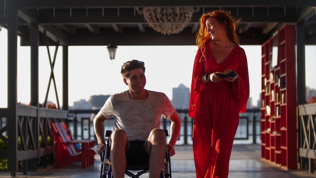 Schönes rothaariges Mädchen rollt abends Mann im Rollstuhl auf der Promenade