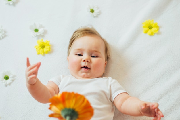 Schönes Säuglingsbaby, das auf weißen Blättern liegt, die für eine Blume greifen.