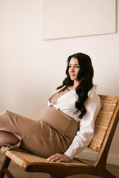 Schönes schwangeres Mädchen mit Locken in einem Kleid in einem hellen Studio mit stilvollem Interieur das Konzept einer glücklichen Schwangerschaft und Familie