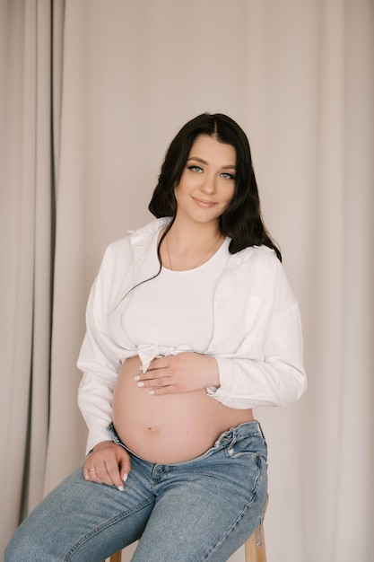 Schönes schwangeres Mädchen mit Locken in weißem Hemd und Jeans in einem hellen Studio mit stilvollem Interieur das Konzept einer glücklichen Schwangerschaft und Familie