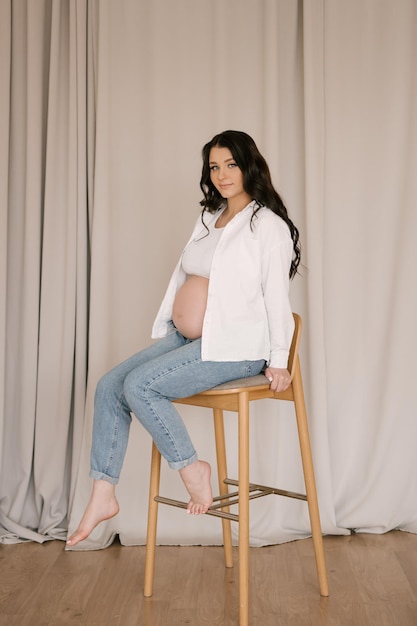 Schönes schwangeres Mädchen mit Locken in weißem Hemd und Jeans in einem hellen Studio mit stilvollem Interieur das Konzept einer glücklichen Schwangerschaft und Familie