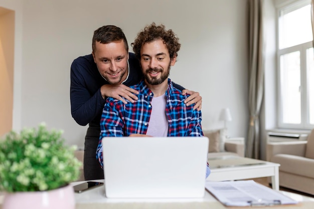 Schönes schwules Paar, das zusammen lacht, während es zu Hause in ihrem Wohnzimmer sitzt Zwei romantische junge männliche Liebhaber, die Spaß haben, drinnen im Internet zu surfen Junges schwules Paar, das zusammen lebt