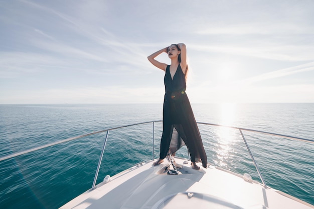 Schönes, selbstbewusstes Mädchen in einem schwarzen Sommerkleid genießt die Seereise auf ihrer Yacht in den Strahlen der Sonne