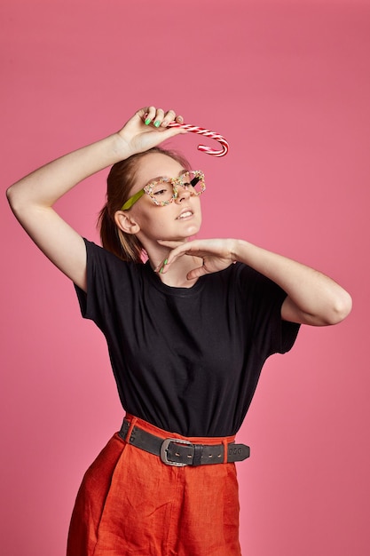 Schönes sexy junges Mädchen in rotem Kleid und süßer Streuselbrille hält eine festliche Zuckerstange in der Hand auf rotem Hintergrund