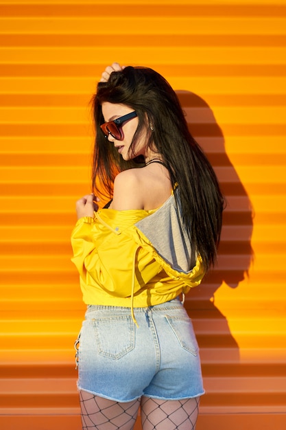 Schönes sexy junges stilvolles Mädchen mit langen brünetten Haaren, die trendige gelbe Jacke, Jeansshorts und modische Sonnenbrille tragen, die auf orange Stadtwand aufwerfen