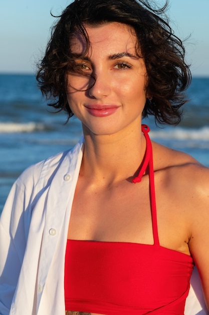 Schönes sexy Mädchen am Meeresstrand in einem roten Kleid betrachtet den flatternden Wind der Kamerahaare bei Sonnenuntergang. Nahaufnahme eines modischen nackten Mädchens in einem Badeanzug. Nahaufnahmemodeporträt des Modells im Freien