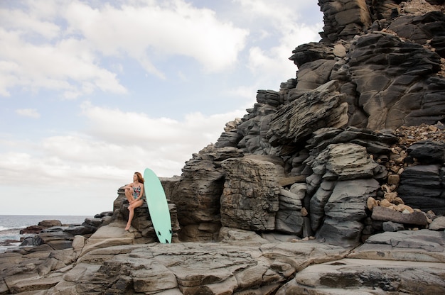 Schönes sexy Mädchen im bunten Badeanzug sitzt in der Nähe des Surfbretts auf dem Felsen über dem Atlantik und dem klaren Himmel