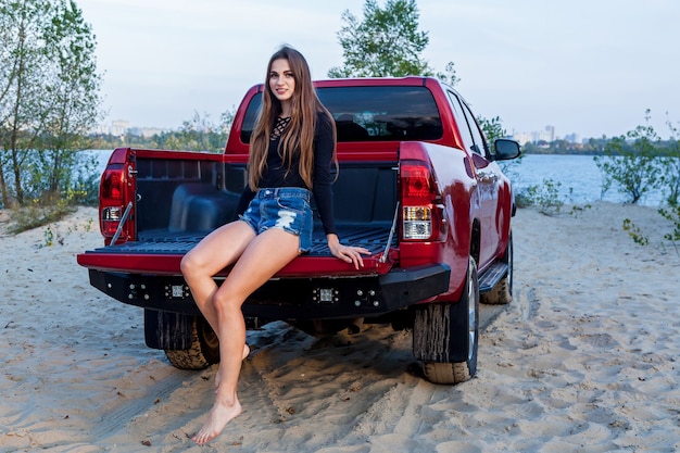 Schönes sexy Mädchen mit langen Haaren in kurzen Jeans-Shorts und einem schwarzen Body posiert in der offenen Karosserie eines roten Pickup-Trucks. Mädchen posiert in der Nähe des Autos