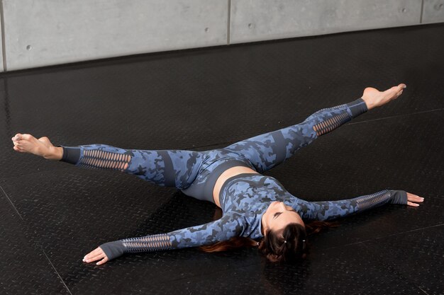 Schönes Sporteignungsmädchen mit Zöpfen. Yoga machen. Liegt auf dem Boden, die Beine in Bindfäden. Tarnblauer Trainingsanzug. Sport und Gesundheit