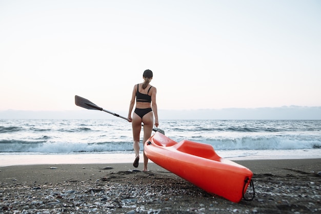 Schönes Sportmädchen in einem schwarzen Badeanzug geht mit einem orangefarbenen Kajak, um bei Sonnenuntergang im Meer zu schwimmen