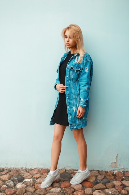 Schönes stilvolles blondes Mädchen in einer Jeansjacke mit Turnschuhen nahe einer blauen Wand am Sommertag