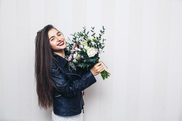 Schönes stilvolles Mädchen in Lederjacke, das einen Blumenstrauß hält und gegen eine weiße Wand posiert