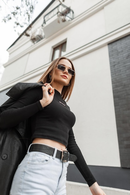 Schönes, stylisches junges Frauenmodell mit Vintage-Sonnenbrille, das ein trendiges schwarzes T-Shirt und Jeans mit einem Mantel trägt, geht in der Stadt in der Nähe des Gebäudes spazieren