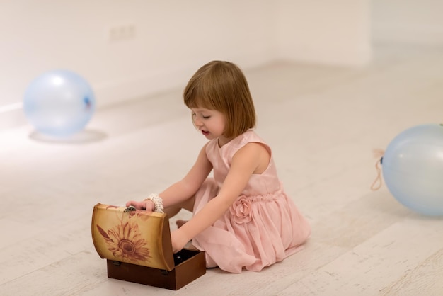 Schönes, süßes kleines Mädchen in rosa Kleid, das es genießt, während es zu Hause mit dem Schmuck der Mutter aus der Schachtel auf dem Boden spielt